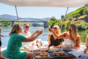 Grupo de turistas brinda com espumante durante um passeio de barco no rio Douro, com paisagem de vinhas e ponte ao fundo – Deltatur.