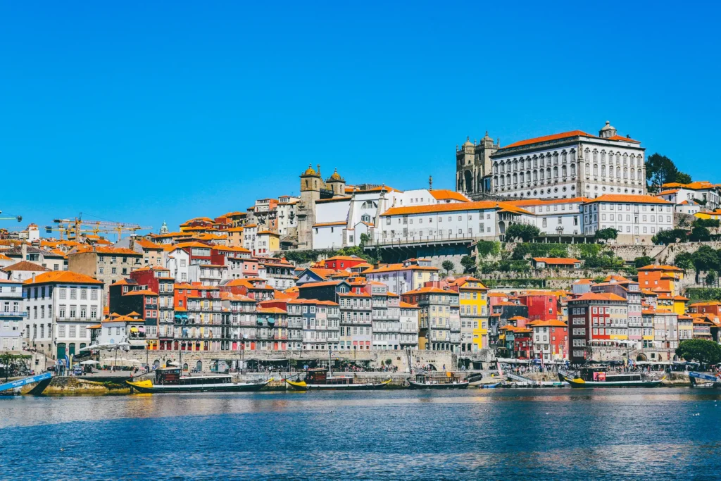 Vista ampla da Ribeira do Porto, com as suas casas coloridas e telhados de terracota, e barcos no Rio Douro sob céu azul.